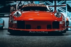 A vibrant red sports car with a sleek and aggressive design is pictured from the front, prominently displaying its headlights and aerodynamic features. The background includes a large Union Jack flag and additional vehicles, indicating an indoor exhibition or showroom setting.