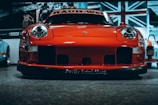 A vibrant red sports car with a sleek and aggressive design is pictured from the front, prominently displaying its headlights and aerodynamic features. The background includes a large Union Jack flag and additional vehicles, indicating an indoor exhibition or showroom setting.