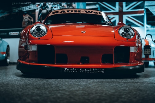 A vibrant red sports car with a sleek and aggressive design is pictured from the front, prominently displaying its headlights and aerodynamic features. The background includes a large Union Jack flag and additional vehicles, indicating an indoor exhibition or showroom setting.
