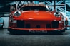 A vibrant red sports car with a sleek and aggressive design is pictured from the front, prominently displaying its headlights and aerodynamic features. The background includes a large Union Jack flag and additional vehicles, indicating an indoor exhibition or showroom setting.