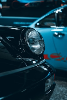 Close-up view of two cars parked side by side, focusing on the headlight of a black vintage car in the foreground. The background features a light blue car with a blurred red logo on its side.