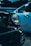 Close-up view of two cars parked side by side, focusing on the headlight of a black vintage car in the foreground. The background features a light blue car with a blurred red logo on its side.