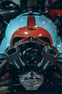 A close-up of a polished, chrome motorcycle engine displaying intricate detailing and mechanical components. The blue and orange vehicle body in the background suggests a vibrant, sporty design.