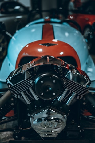 A close-up of a polished, chrome motorcycle engine displaying intricate detailing and mechanical components. The blue and orange vehicle body in the background suggests a vibrant, sporty design.