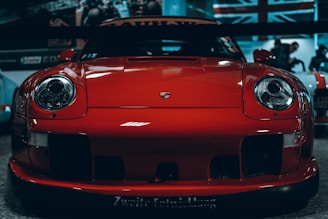 A bright red sports car is prominently displayed in a showroom or garage setting. The car features a sleek design with large, distinctive round headlights and a low, aerodynamic body. The Porsche emblem is visible on the hood. In the background, blurred images include a British flag and other cars.