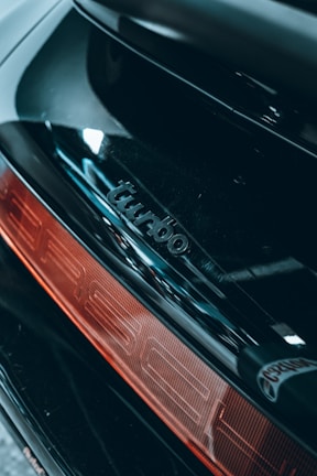 Close-up of a car's rear section featuring a shiny black surface with the word 'turbo' embossed above a red taillight.