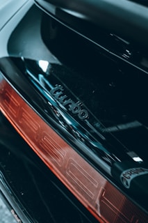 Close-up of a car's rear section featuring a shiny black surface with the word 'turbo' embossed above a red taillight.