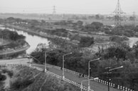 A winding road bordered by black and white striped barriers curves alongside a narrow river or canal, with dense foliage and power lines seen in the background. A tall transmission tower stands prominently to the right, and scattered street lamps line the road. The landscape appears expansive and somewhat rural.