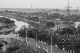 A winding road bordered by black and white striped barriers curves alongside a narrow river or canal, with dense foliage and power lines seen in the background. A tall transmission tower stands prominently to the right, and scattered street lamps line the road. The landscape appears expansive and somewhat rural.