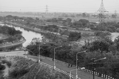 A winding road bordered by black and white striped barriers curves alongside a narrow river or canal, with dense foliage and power lines seen in the background. A tall transmission tower stands prominently to the right, and scattered street lamps line the road. The landscape appears expansive and somewhat rural.