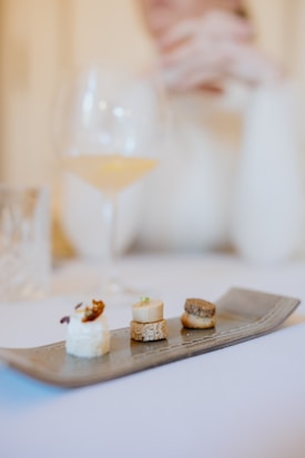 A sleek, modern platter displays an assortment of three elegant bite-sized appetizers. Each piece is crafted with precise attention to detail, layered beautifully on small rounds of bread. In the background, a blurry figure seems to be seated, accompanied by a glass of white wine and a decanter.