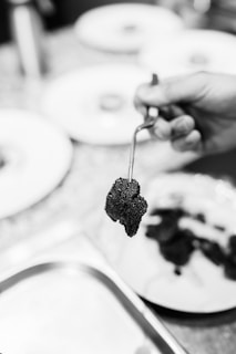 Hand holding a kitchen utensil with a piece of food, possibly a truffle, in focus. The background is slightly blurred, showing plates and other cooking activities.