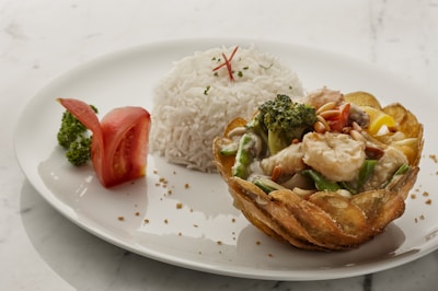 A white plate features a beautifully arranged dish with a serving of white rice and a separate potato crusted basket filled with vegetables such as broccoli and bell peppers, as well as pieces of seafood and pine nuts. A slice of tomato is also placed on the plate.