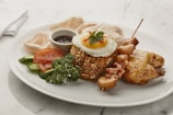 A dish featuring a mound of spiced rice topped with a sunny side up egg, garnished with a small parsley leaf and red chili slices. It is served alongside grilled chicken satay skewers, a piece of fried chicken, cucumber slices, tomato slices, prawn crackers, and a sprig of parsley. A small dish of dark sauce accompanies the meal on a white plate, set on a marble surface.