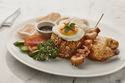 A dish featuring a mound of spiced rice topped with a sunny side up egg, garnished with a small parsley leaf and red chili slices. It is served alongside grilled chicken satay skewers, a piece of fried chicken, cucumber slices, tomato slices, prawn crackers, and a sprig of parsley. A small dish of dark sauce accompanies the meal on a white plate, set on a marble surface.