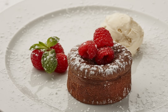 A chocolate dessert dusted with powdered sugar sits beautifully on a white plate. It is topped with vibrant red raspberries and accompanied by a dollop of vanilla ice cream. Fresh mint leaves and additional raspberries are placed beside it, adding a touch of color and freshness.