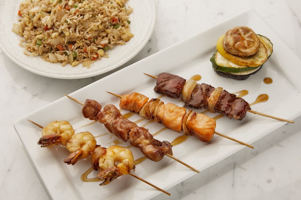 a white plate topped with skewers of food next to a bowl of rice