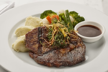 A well-cooked steak garnished with herbs is placed on a white plate, accompanied by mashed potatoes and assorted steamed vegetables including broccoli, carrots, and green beans. A small bowl of sauce is set beside the steak.
