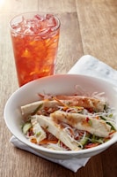 A bowl filled with vermicelli noodles topped with sliced grilled chicken, cucumber, carrot, and green onion. The dish is garnished with herbs and a light sauce. Next to the bowl, there is a glass of iced tea with ice cubes on a wooden table, accompanied by a napkin nearby.