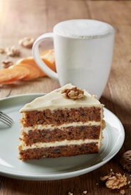 A slice of layered carrot cake with cream cheese frosting on a plate, garnished with a walnut on top. A white cup filled with a frothy beverage sits in the background on a wooden surface, accompanied by scattered walnuts and a slice of dried apricot.