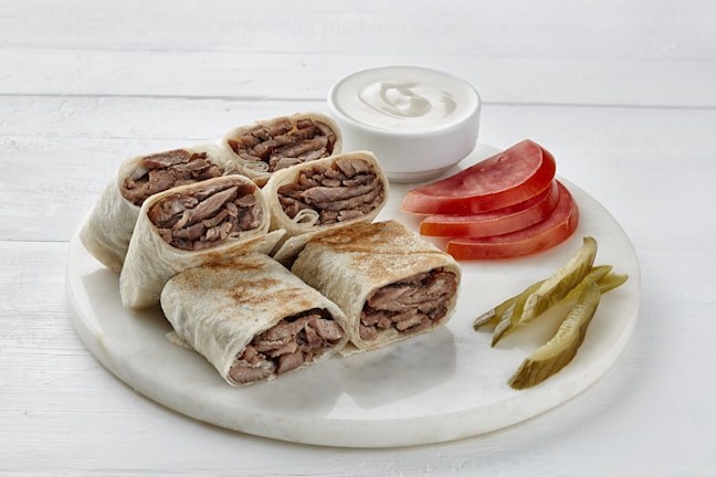 A plate of grilled beef wraps cut into half, served alongside slices of fresh tomato and pickles. A small bowl of white sauce accompanies the dish, which is presented on a light, neutral background.