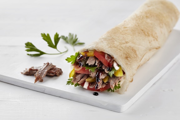A neatly arranged wrap placed on a white rectangular board. The wrap is filled with colorful ingredients including lettuce, tomatoes, yellow bell peppers, and some meat, with a garnish of parsley on the side. A small piece of meat and a sprig of parsley rest beside the wrap on the board.