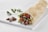 A neatly arranged wrap placed on a white rectangular board. The wrap is filled with colorful ingredients including lettuce, tomatoes, yellow bell peppers, and some meat, with a garnish of parsley on the side. A small piece of meat and a sprig of parsley rest beside the wrap on the board.