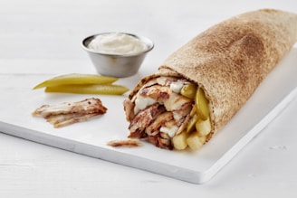 A close-up view of a rolled sandwich, likely a shawarma, on a light wooden board. The sandwich consists of grilled meat slices, pickles, and sauces wrapped in thin flatbread. Beside it, there are slices of pickles and a small metal cup filled with a creamy white sauce.
