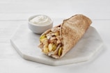 A pita wrap filled with slices of meat, pickles, and sauce, placed on a white hexagonal plate with a small bowl of white creamy sauce beside it.