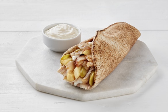 A pita wrap filled with slices of meat, pickles, and sauce, placed on a white hexagonal plate with a small bowl of white creamy sauce beside it.