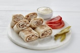 Several rolled flatbreads filled with sliced chicken are placed on a white round serving tray. Accompanying them are sliced tomatoes, pickles, and a small bowl of creamy sauce.