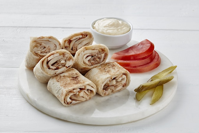 Several rolled flatbreads filled with sliced chicken are placed on a white round serving tray. Accompanying them are sliced tomatoes, pickles, and a small bowl of creamy sauce.