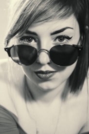 A monochrome close-up portrait of an individual wearing dark sunglasses and looking directly at the camera. The person has light hair styled with a side-swept fringe, and their expression appears neutral. The lighting and focus create a vintage or classic feel to the image.