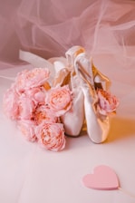 A pair of elegant ballet shoes is positioned in a graceful manner next to a bouquet of delicate, pale pink flowers. A soft, heart-shaped decoration lies nearby on a sheer, neutral-toned fabric background.