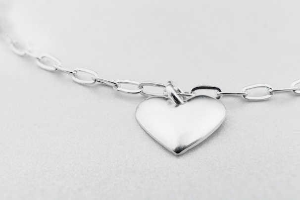 A heart-shaped silver pendant attached to a glossy chain with oval links, set against a soft, light gray background. The whole setup gives a minimalist and elegant impression.