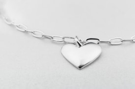 A heart-shaped silver pendant attached to a glossy chain with oval links, set against a soft, light gray background. The whole setup gives a minimalist and elegant impression.