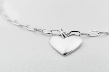 A heart-shaped silver pendant attached to a glossy chain with oval links, set against a soft, light gray background. The whole setup gives a minimalist and elegant impression.