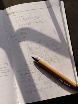 A page from a calculus textbook is open, showing several mathematical equations and formulas. A pen with a wooden body is placed diagonally across the page, casting a shadow over the lower section of the book. The lighting suggests a natural source, possibly sunlight, coming from the upper left corner.