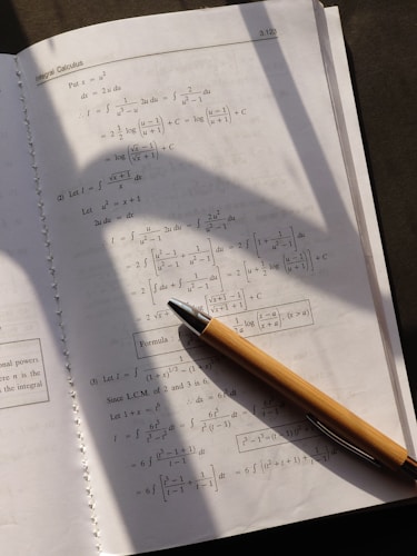A page from a calculus textbook is open, showing several mathematical equations and formulas. A pen with a wooden body is placed diagonally across the page, casting a shadow over the lower section of the book. The lighting suggests a natural source, possibly sunlight, coming from the upper left corner.