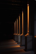 A series of illuminated columns casting warm, orange light in a dimly lit area, creating a dramatic interplay of light and shadows on the ground. The architectural design features tall, vertical elements with a classic style, each column highlighted by the focused lighting.