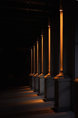 A series of illuminated columns casting warm, orange light in a dimly lit area, creating a dramatic interplay of light and shadows on the ground. The architectural design features tall, vertical elements with a classic style, each column highlighted by the focused lighting.