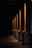 A series of illuminated columns casting warm, orange light in a dimly lit area, creating a dramatic interplay of light and shadows on the ground. The architectural design features tall, vertical elements with a classic style, each column highlighted by the focused lighting.