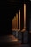 A series of illuminated columns casting warm, orange light in a dimly lit area, creating a dramatic interplay of light and shadows on the ground. The architectural design features tall, vertical elements with a classic style, each column highlighted by the focused lighting.
