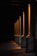 A series of illuminated columns casting warm, orange light in a dimly lit area, creating a dramatic interplay of light and shadows on the ground. The architectural design features tall, vertical elements with a classic style, each column highlighted by the focused lighting.