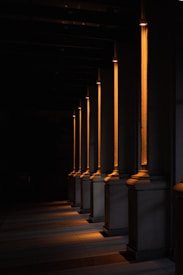 A series of illuminated columns casting warm, orange light in a dimly lit area, creating a dramatic interplay of light and shadows on the ground. The architectural design features tall, vertical elements with a classic style, each column highlighted by the focused lighting.