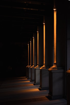 A series of illuminated columns casting warm, orange light in a dimly lit area, creating a dramatic interplay of light and shadows on the ground. The architectural design features tall, vertical elements with a classic style, each column highlighted by the focused lighting.