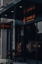 An exterior view of a coffee shop named 'Charlie Coffee & Brew' featuring dark, modern signage with a warm glow. The shop has large glass windows reflecting exterior light and a set of outdoor chairs and a table made of wood, creating a cozy and inviting atmosphere.