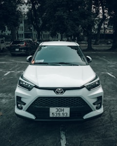 A white Toyota car is parked in an outdoor parking lot. The vehicle is positioned straight, facing forward, and surrounded by several large trees. The scene appears overcast, creating a muted light effect.