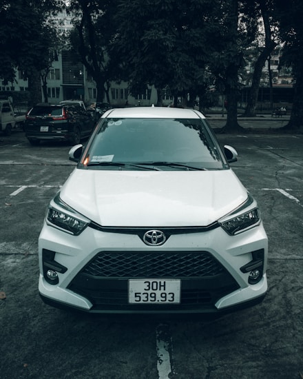A white Toyota car is parked in an outdoor parking lot. The vehicle is positioned straight, facing forward, and surrounded by several large trees. The scene appears overcast, creating a muted light effect.