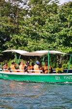A group of people wearing orange life vests are seated in a green motorboat with a white canopy. The boat is on a body of water and is surrounded by lush green trees. Some of them appear to be enjoying the ride, and there is text on the boat's hull that reads 'BY FAITH.'